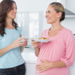 Happy pregnant woman holding cookies and her friend in the kitchen